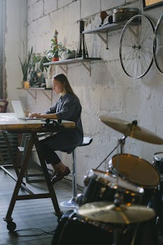 Side view of focused female freelancer wearing casual clothes sitting and typing on modern laptop in creative room with drums